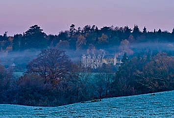 Historic country estate partially veiled in morning mist, surrounded by autumn trees and rolling frosted fields beneath a soft pink sunrise sky.