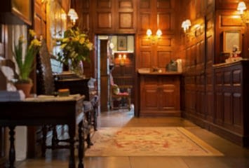 Warm wood-panelled reception area at Gravetye Manor, featuring antique furnishings, soft lighting, floral arrangements, and a traditional rug leading into an adjoining room.