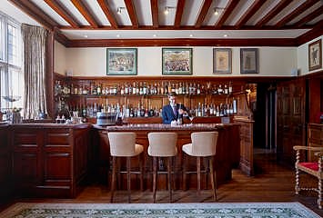 Elegant wooden bar at Gravetye Manor with a beamed ceiling, extensive spirits display, framed artwork, and a bartender preparing drinks behind three upholstered bar stools.