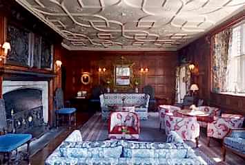 Historic wood-paneled sitting room with ornate ceiling, patterned sofas, armchairs, and a large decorative fireplace.