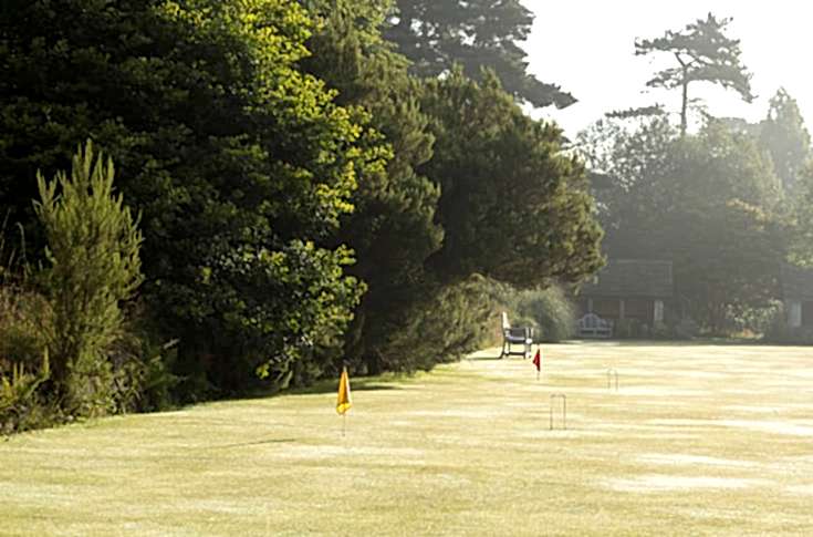 A sunny dewy morning at Gravetye Manor's croquet grounds, large green bushes and trees line the playing area.