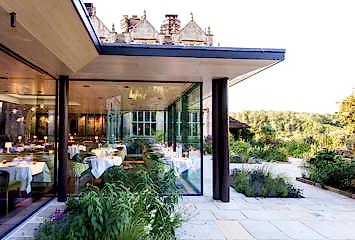 Glass-fronted Michelin-starred restaurant at Gravetye Manor, overlooking landscaped gardens views, with elegant table settings visible through the modern design.