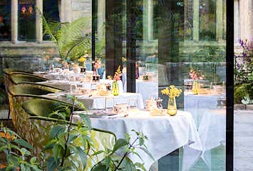 Elegant restaurant tables set with white linens, glassware, and small floral arrangements inside a glass-walled dining room overlooking lush greenery at Gravetye Manor