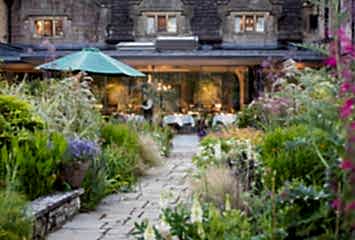 Lush garden walkway leading to a glass-fronted restaurant terrace with outdoor seating, surrounded by colorful flowers and greenery at a historic stone building.