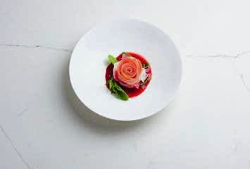 Fine-dining dish presented as a delicate rose-shaped arrangement of thinly sliced vegetables on a white plate, served with a vibrant red sauce and fresh greens.