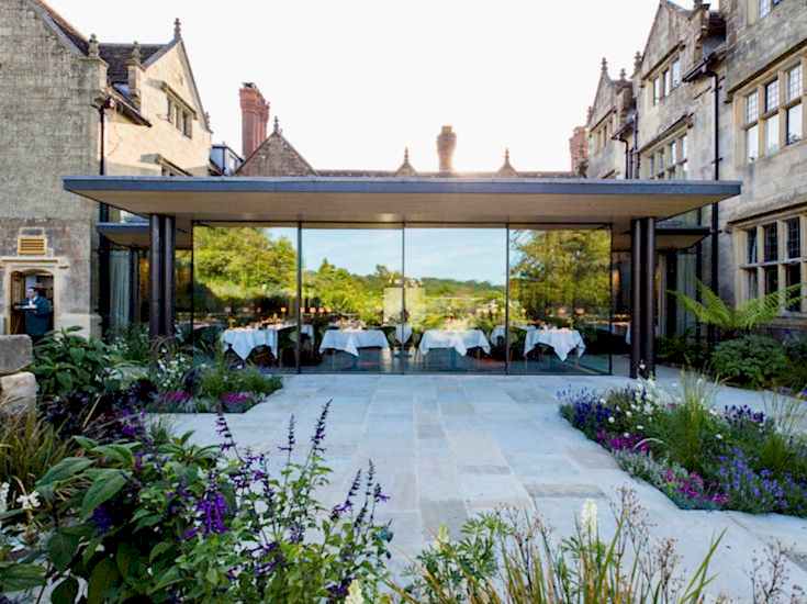 Exterior view of the glass-walled dining room at Gravetye Manor in West Sussex, overlooking landscaped gardens and countryside views.
